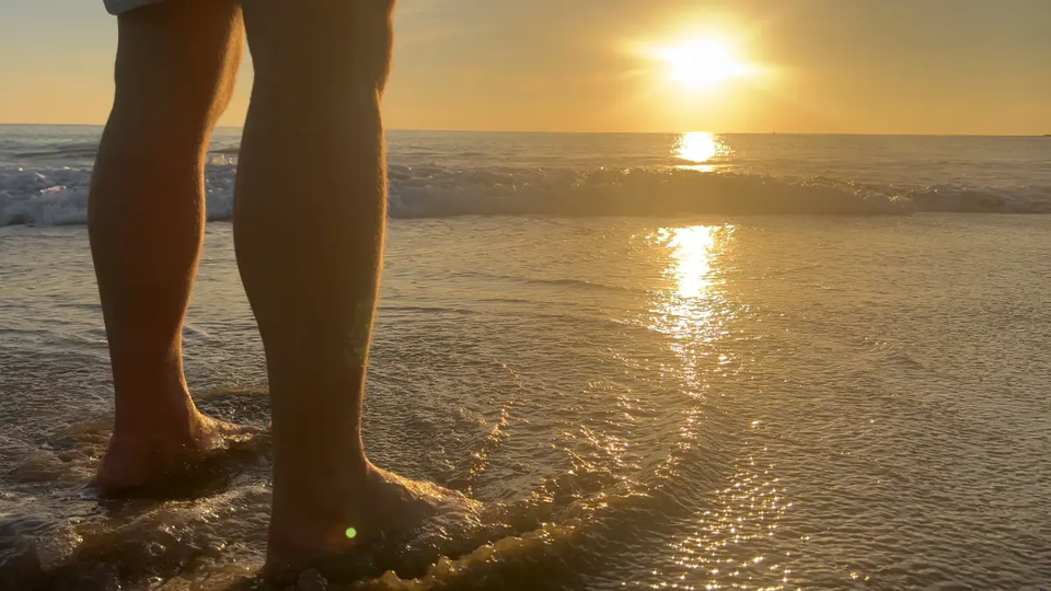 Bare feet standing in shallow ocean water at sunset, with warm sunlight reflecting across the waves, representing natural recovery and connection with nature.