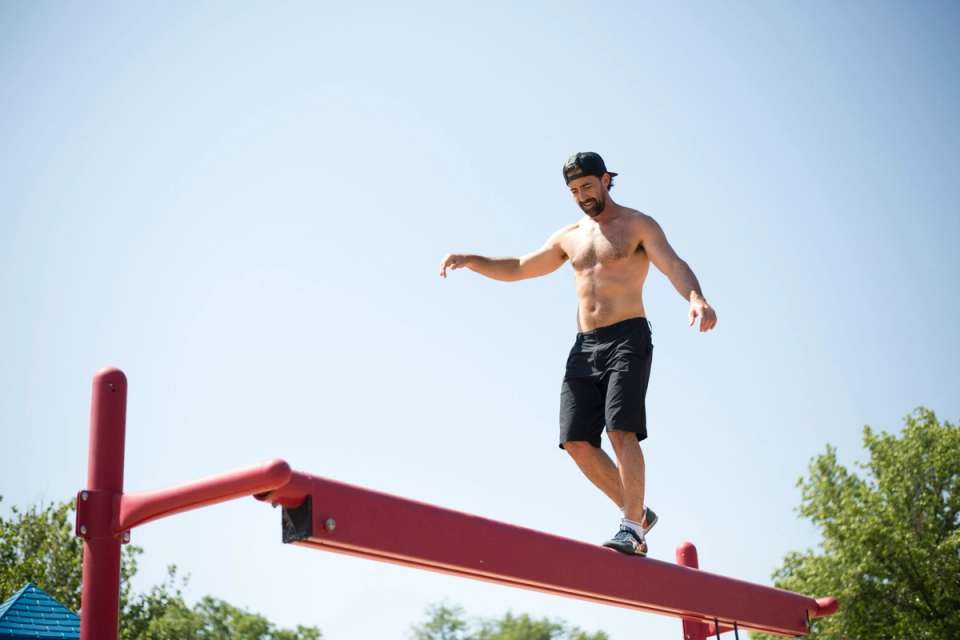 Shirtless man balancing on a narrow outdoor beam with arms extended for stability, demonstrating coordination, proprioception, and full-body control in a natural movement environment.