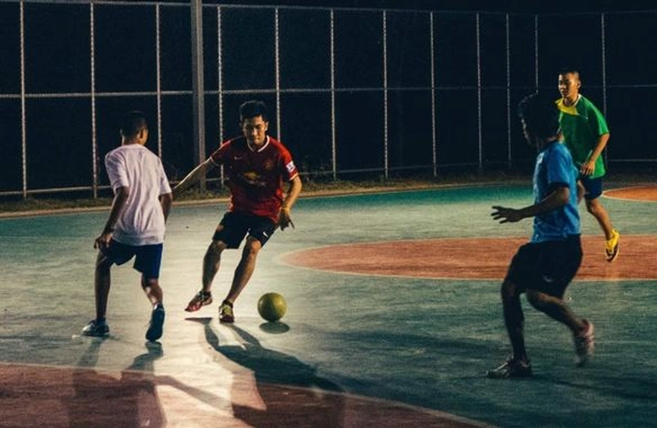Four young footballers play a fast-paced small-sided match on an outdoor hard court at night under floodlights, with one player dribbling as defenders close in.