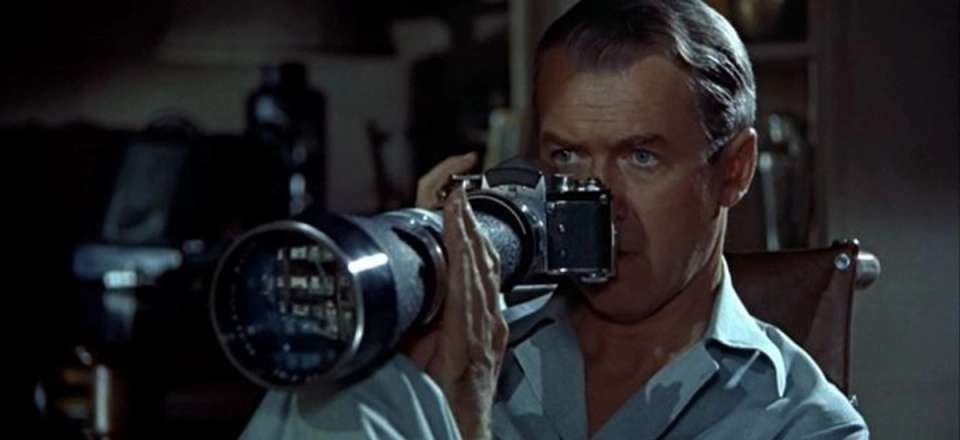 A man peers through a camera with a large telephoto lens, illustrating the act of closely observing or measuring a subject from a distance.