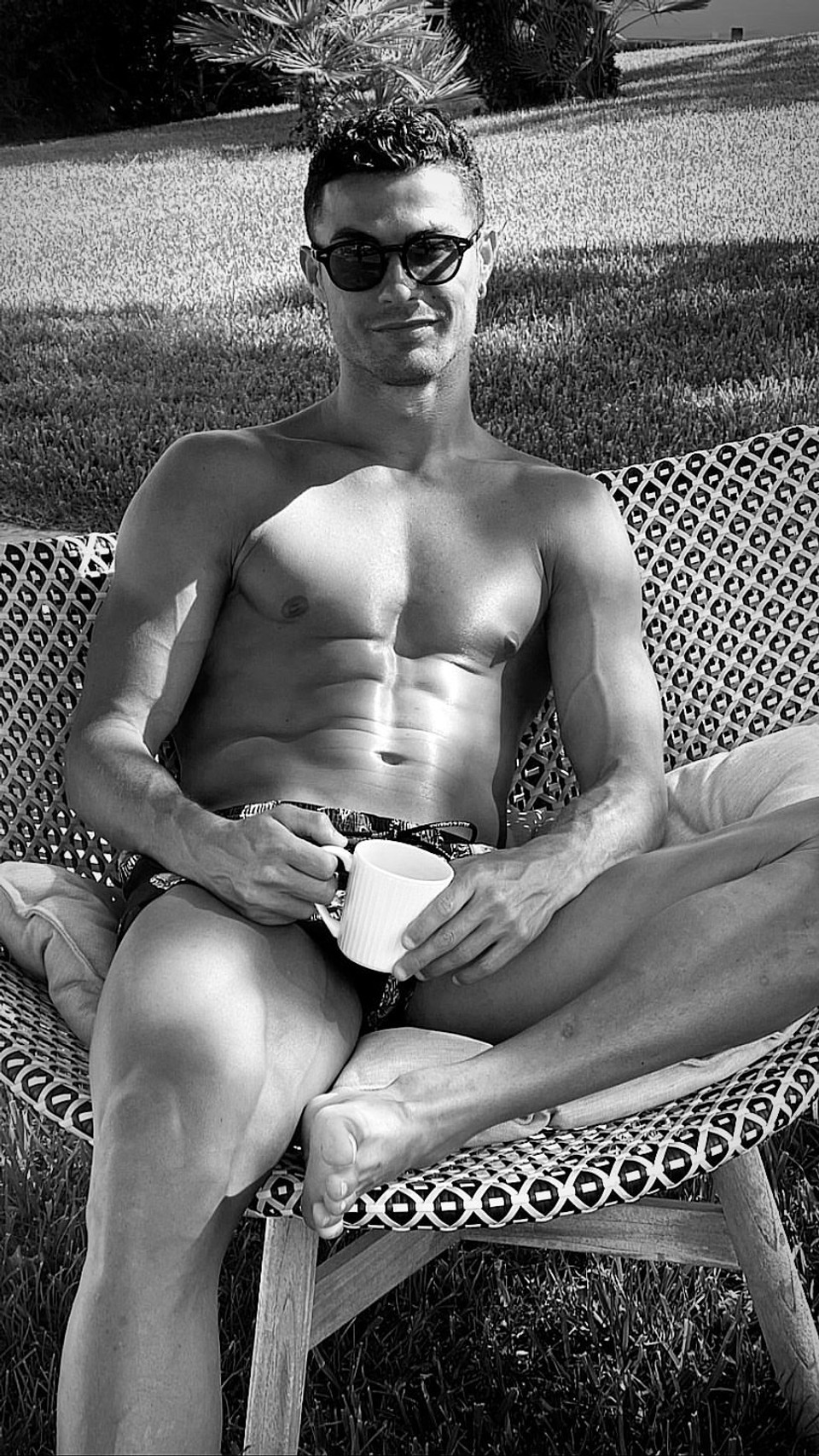 Cristiano Ronaldo sits shirtless outdoors on a cushioned lounge chair holding a mug, illustrating rest and recovery during downtime away from training.