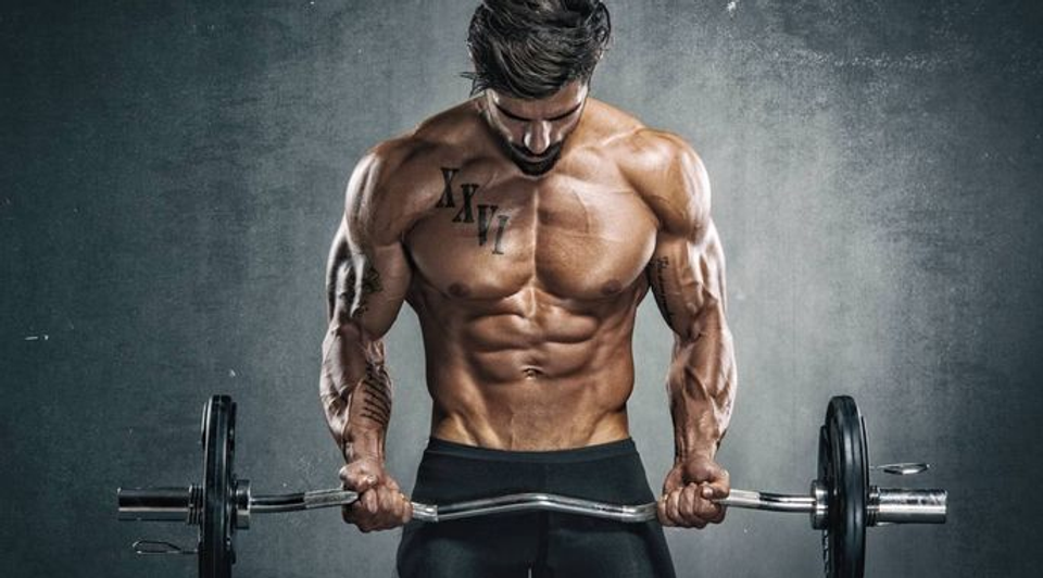 Muscular man performing a barbell curl in a gym, representing the bodybuilding approach contrasted with the natural development of world-class footballers.