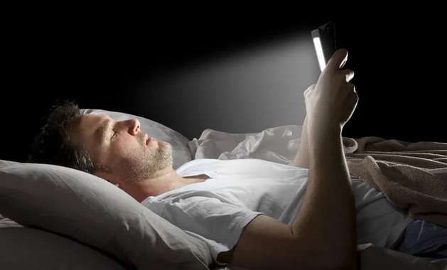 A man lying in bed in the dark, his face lit by the phone he holds above him.