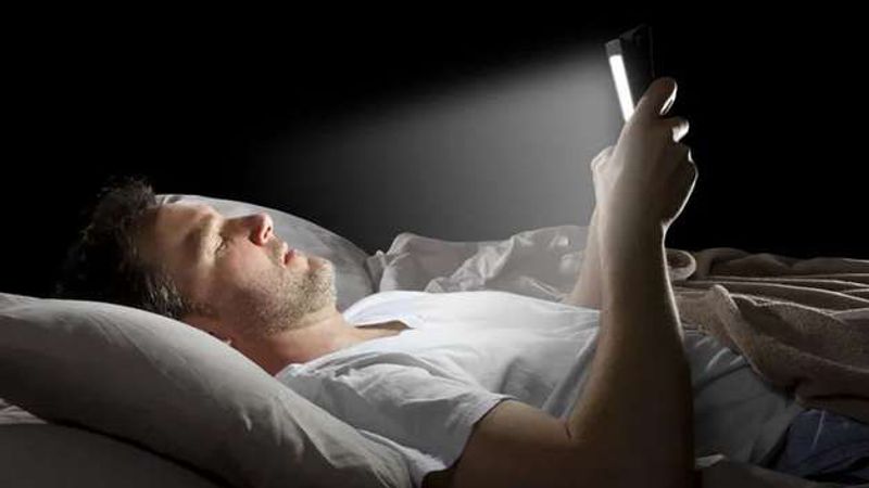 A man lying in bed in the dark, his face lit by the phone he holds above him.