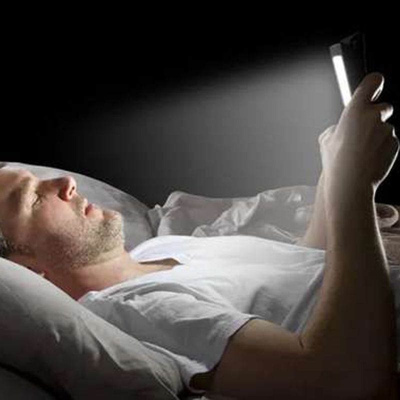 A man lying in bed in the dark, his face lit by the phone he holds above him.