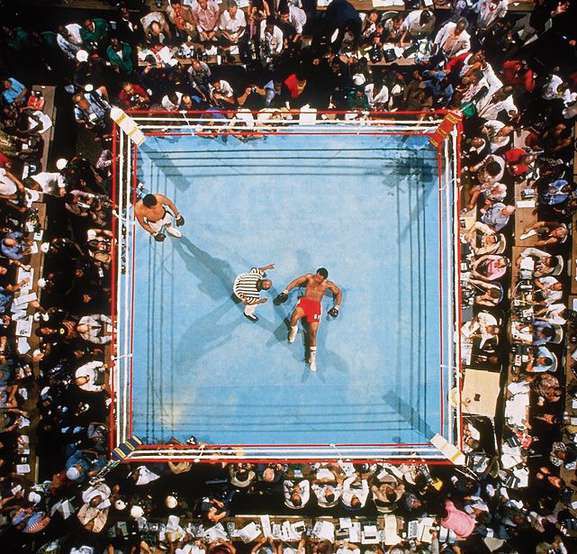 A boxing ring seen from above, surrounded by spectators, a fighter lying on the canvas as the referee stands over him.