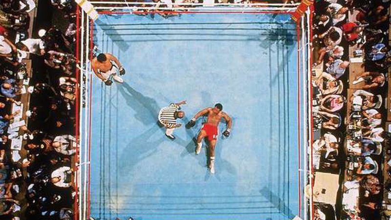 A boxing ring seen from above, surrounded by spectators, a fighter lying on the canvas as the referee stands over him.