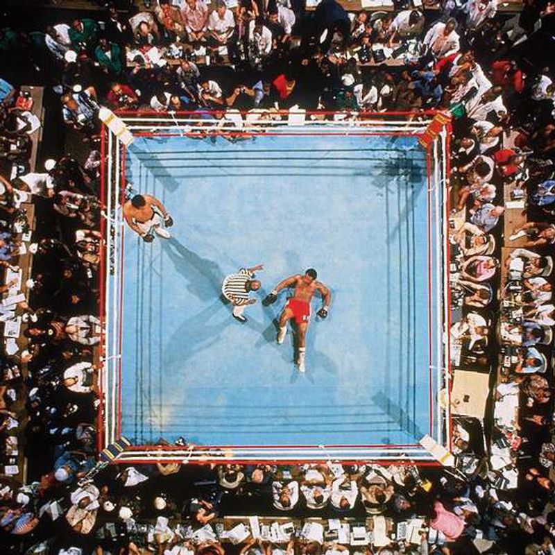 A boxing ring seen from above, surrounded by spectators, a fighter lying on the canvas as the referee stands over him.
