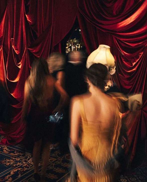 Women in evening dresses, blurred in motion, pass through heavy red velvet curtains in a dim club.