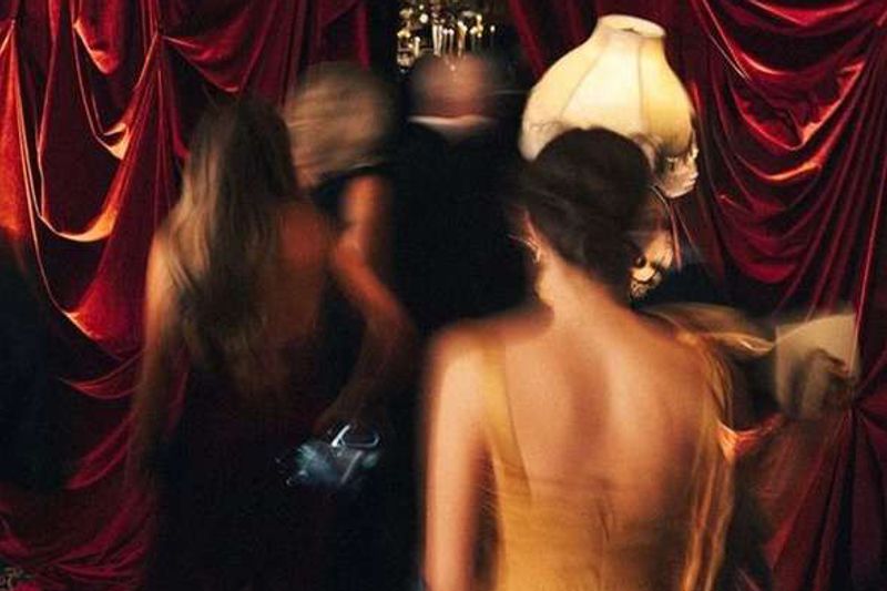Women in evening dresses, blurred in motion, pass through heavy red velvet curtains in a dim club.
