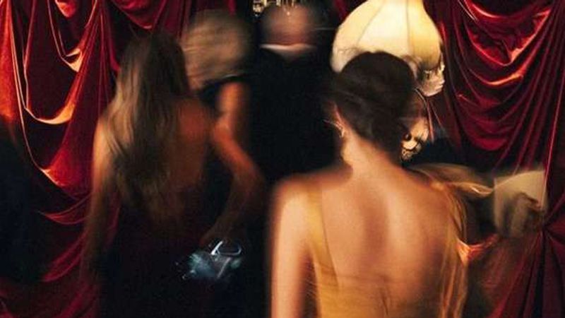 Women in evening dresses, blurred in motion, pass through heavy red velvet curtains in a dim club.