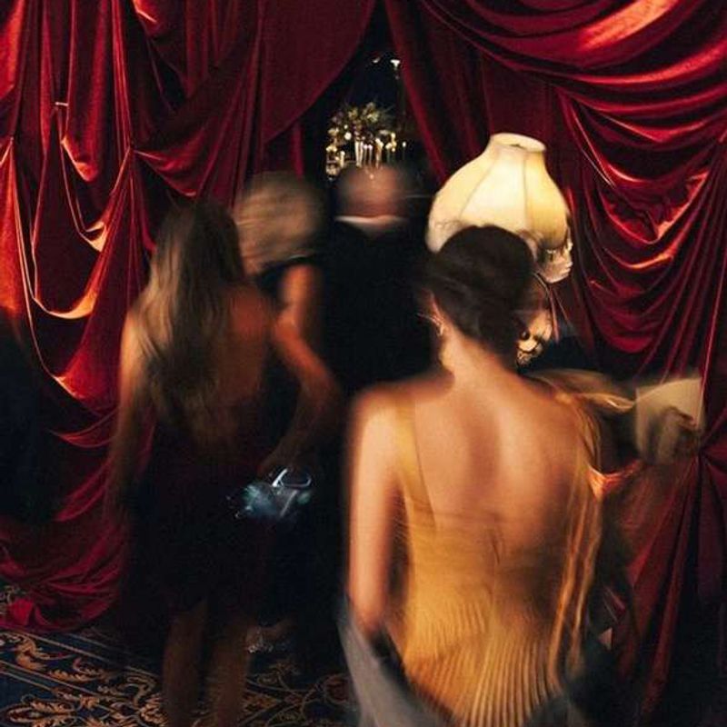 Women in evening dresses, blurred in motion, pass through heavy red velvet curtains in a dim club.