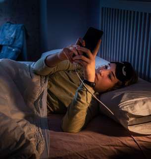 A young woman lying in bed at night, looking at her phone as it charges.