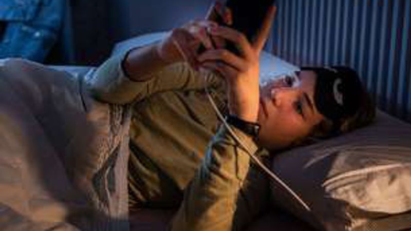 A young woman lying in bed at night, looking at her phone as it charges.