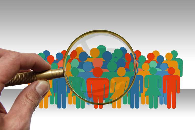 A hand holds a magnifying glass over a crowd of colorful cut-out figures of people.