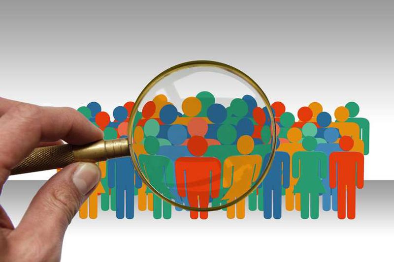 A hand holds a magnifying glass over a crowd of colorful cut-out figures of people.