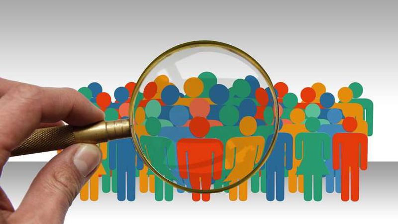 A hand holds a magnifying glass over a crowd of colorful cut-out figures of people.