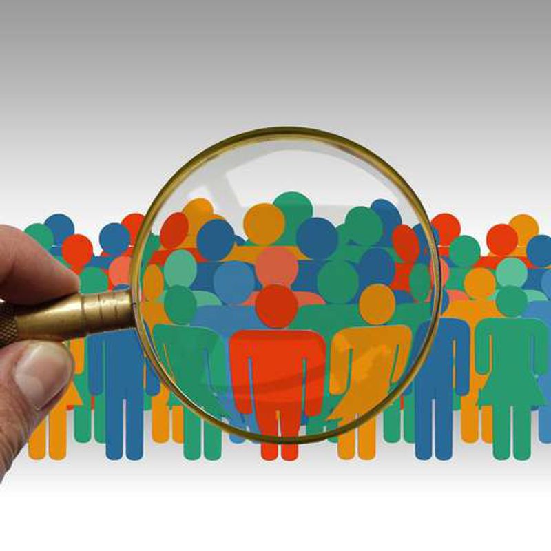 A hand holds a magnifying glass over a crowd of colorful cut-out figures of people.
