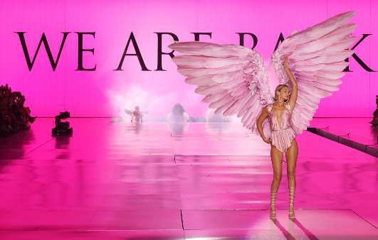 Gigi Hadid on a bright pink runway wearing large pink feathered wings, under the words “We are back”.