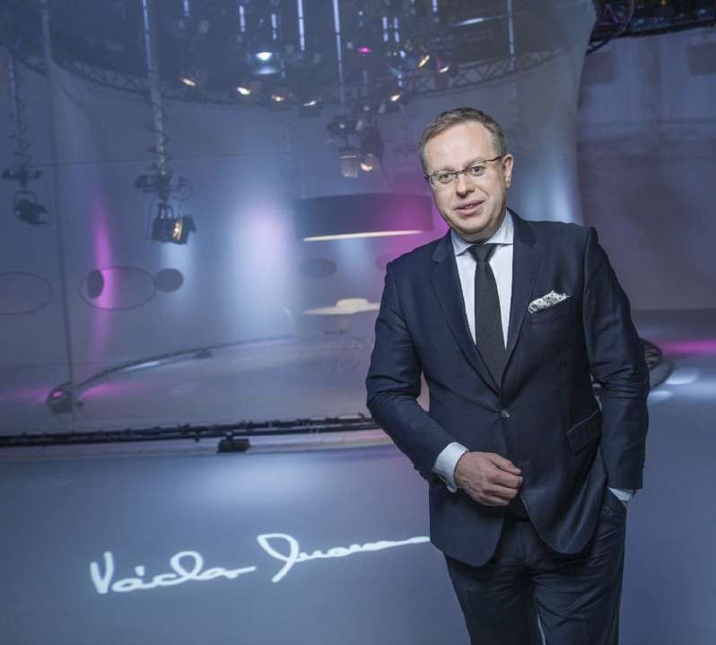 Václav Moravec in a dark suit in an empty television studio, his signature projected on the floor.
