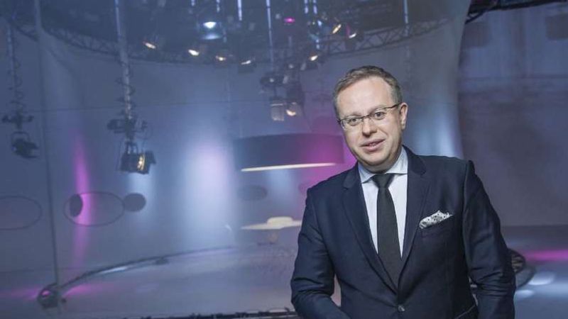 Václav Moravec in a dark suit in an empty television studio, his signature projected on the floor.