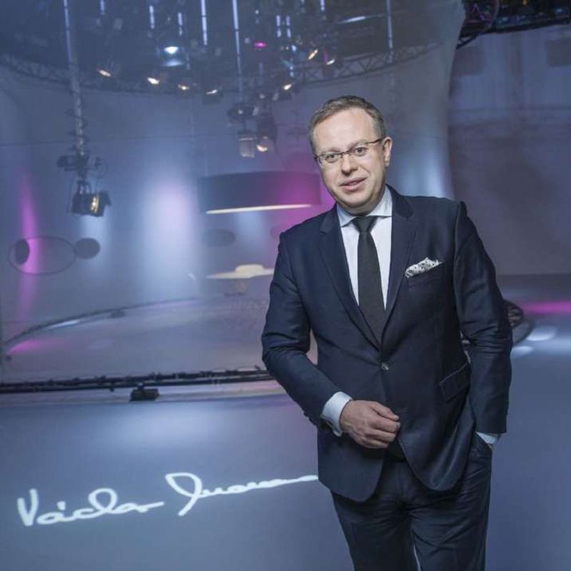 Václav Moravec in a dark suit in an empty television studio, his signature projected on the floor.