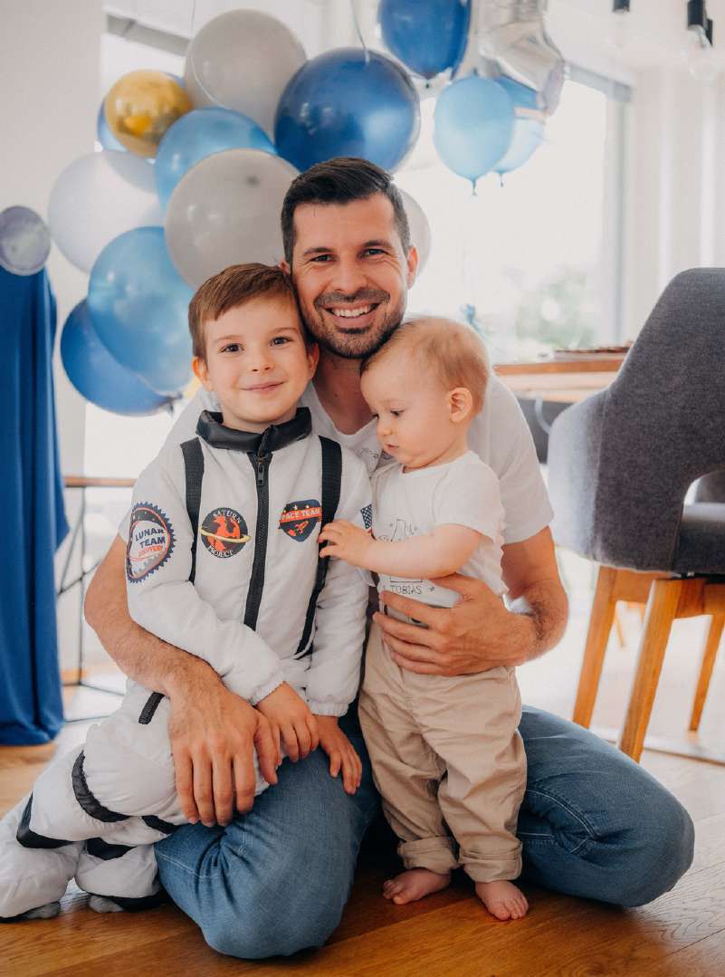 Stanislav Gálik kneels, smiling, with his two young sons, one in an astronaut costume, in front of blue and silver balloons.