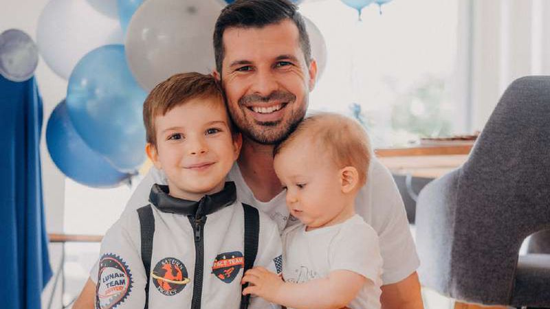 Stanislav Gálik kneels, smiling, with his two young sons, one in an astronaut costume, in front of blue and silver balloons.