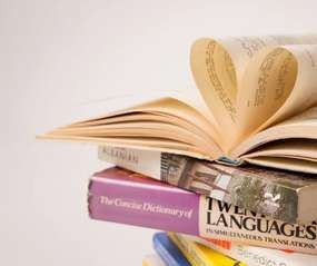 An open book with pages curled into a heart, lying on a stack of language dictionaries.