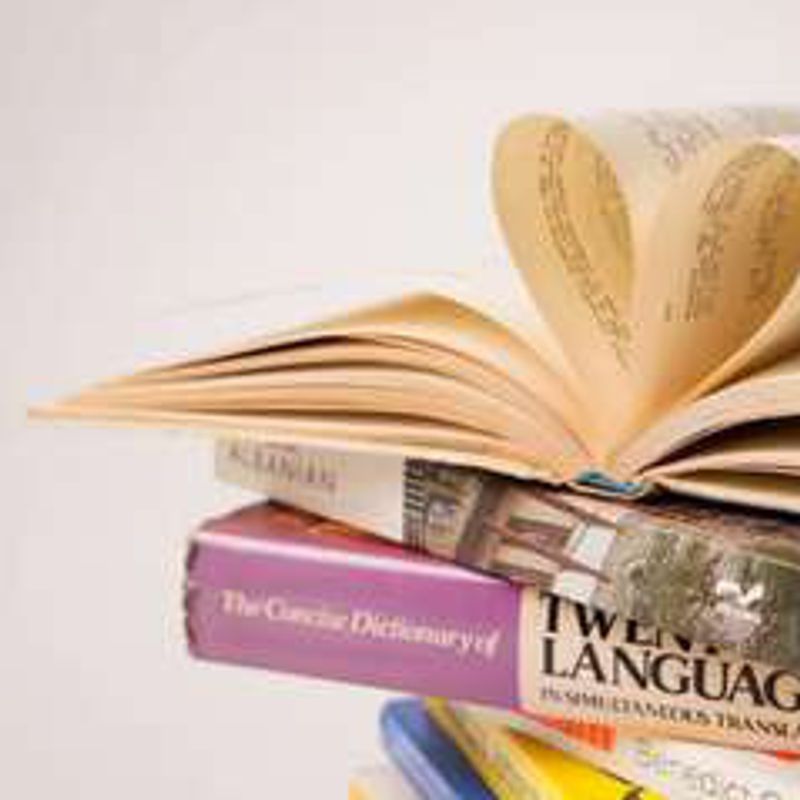An open book with pages curled into a heart, lying on a stack of language dictionaries.