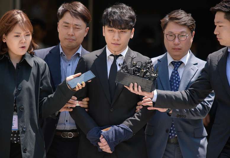 Seungri, in a dark suit, is escorted through a crowd of reporters holding out microphones.