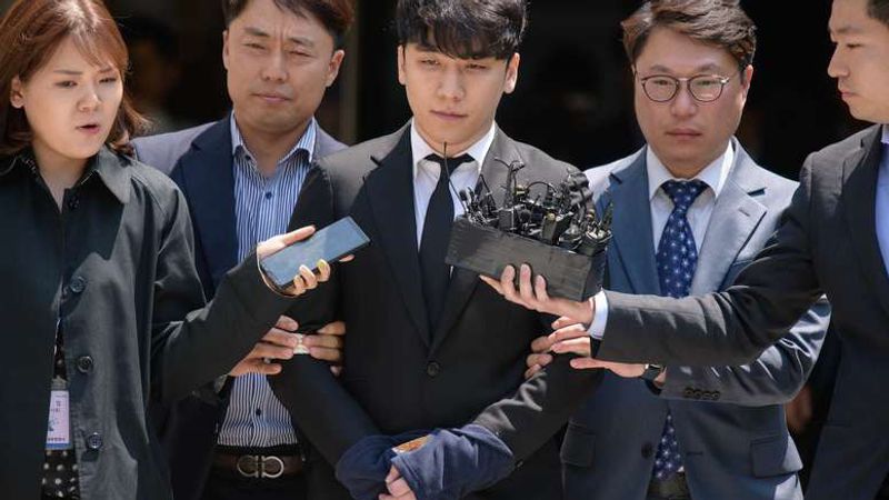 Seungri, in a dark suit, is escorted through a crowd of reporters holding out microphones.