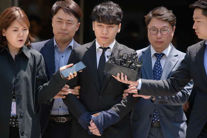 Seungri, in a dark suit, is escorted through a crowd of reporters holding out microphones.