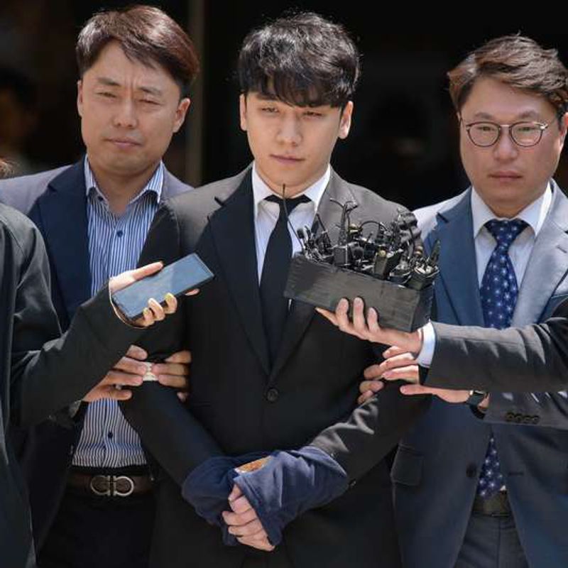 Seungri, in a dark suit, is escorted through a crowd of reporters holding out microphones.