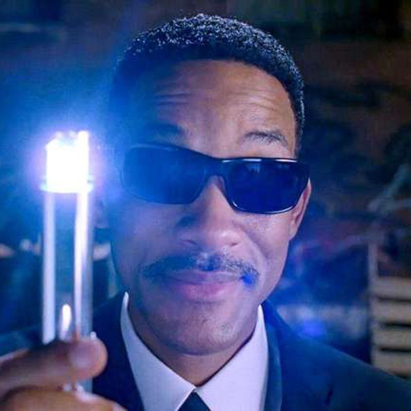 Will Smith in black sunglasses holds up a glowing neuralyzer.