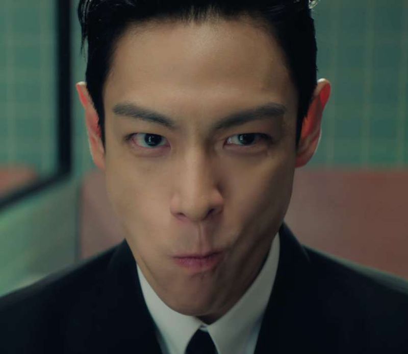 Close-up of T.O.P in a dark suit, looking into the camera with his cheeks puffed out.