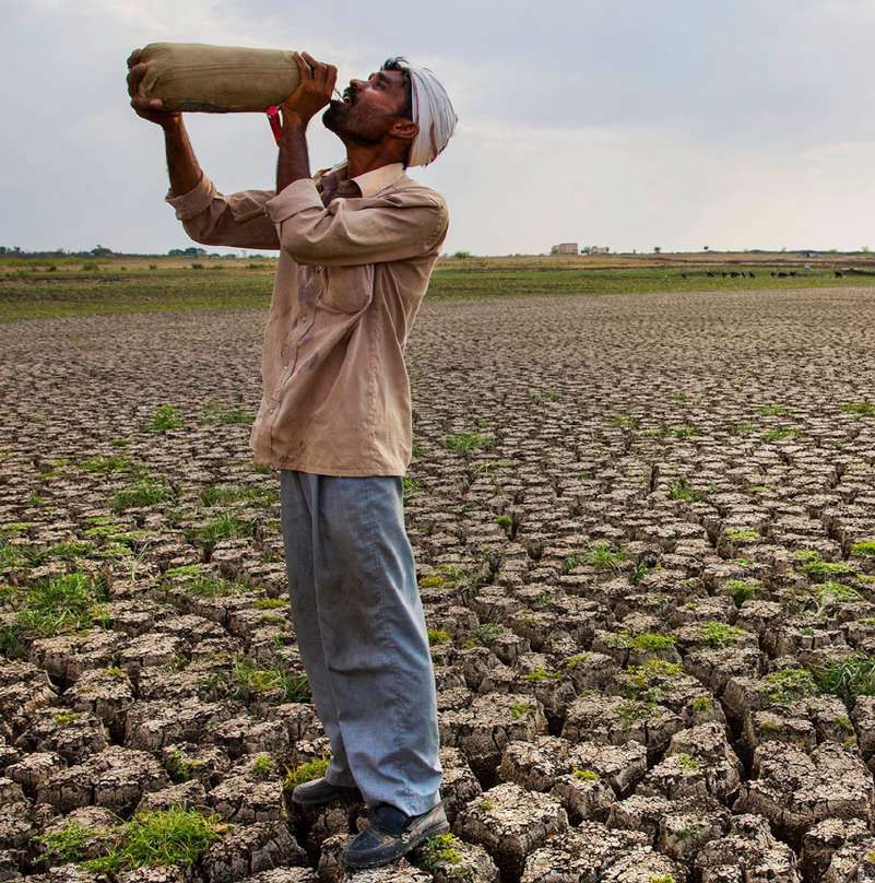 A man in a turban drinks from a water container while standing on the cracked, dry bed of a reservoir.