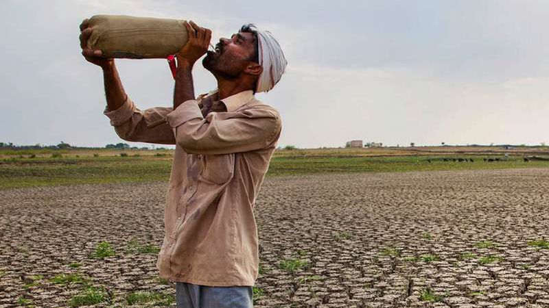 A man in a turban drinks from a water container while standing on the cracked, dry bed of a reservoir.