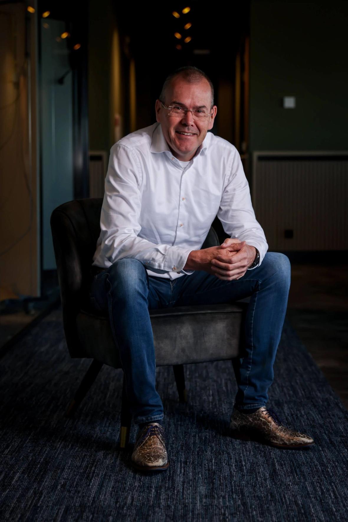 Roel Van Stokkum Eigenaar Elektrobedrijf Verweij in een wit overhemd en spijkerbroek zit op een stoel in een schemerige gang en lacht naar de camera.
