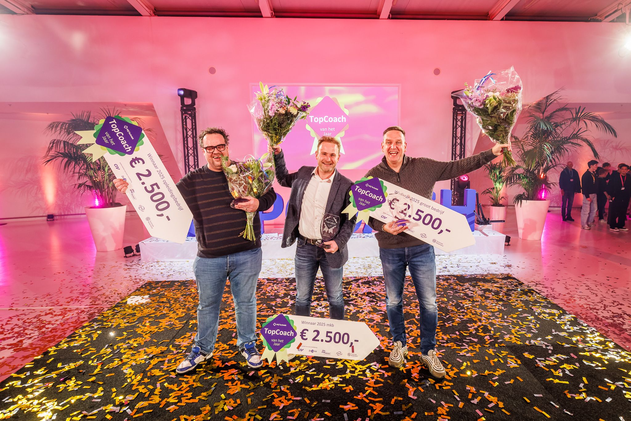 Drie lachende winnaars Topcoach 2024 op podium met award en geldprijs in de hand.