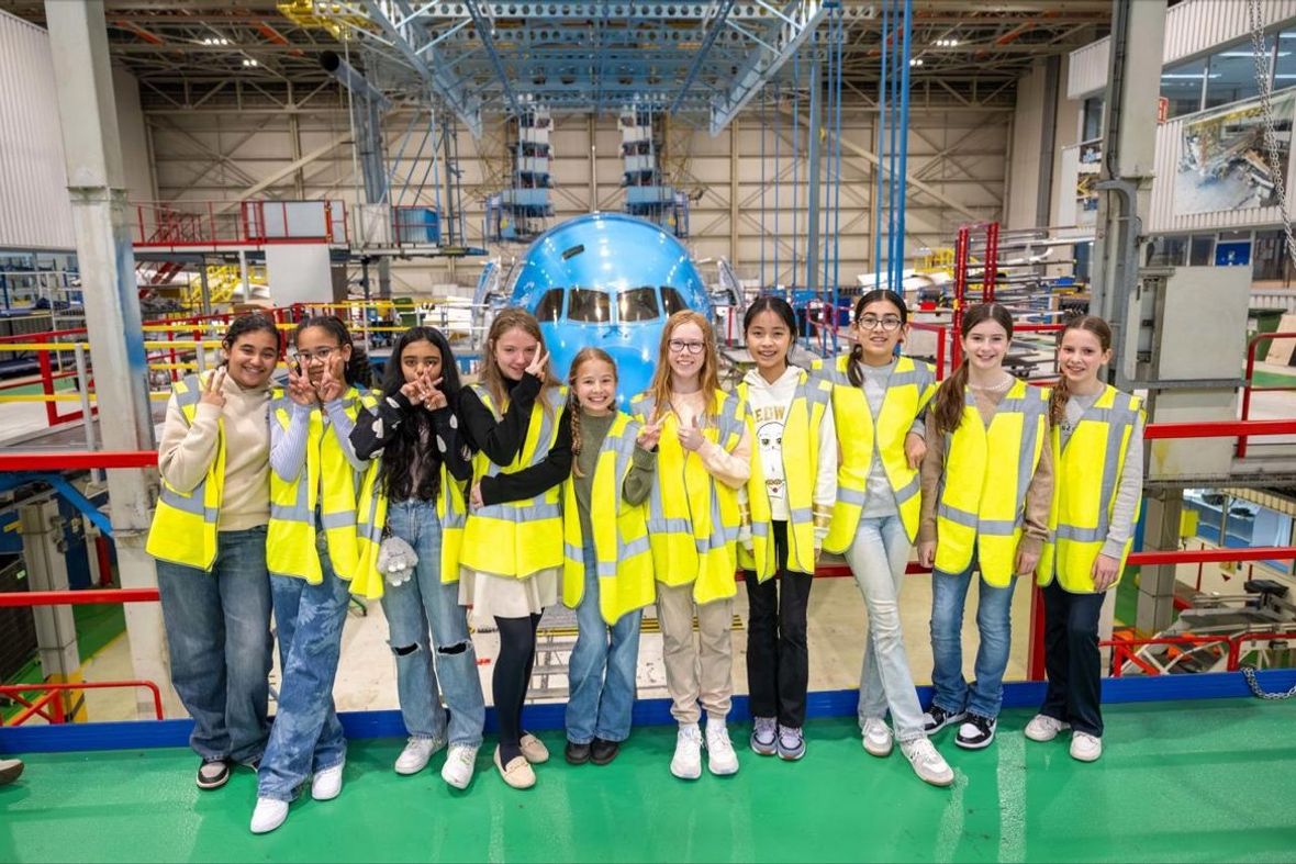 Een groep van tien jonge meiden met gele veiligheidsvesten en vrijetijdskleding staat op een rij in een industriële omgeving met een KLM vliegtuig op de achtergrond.