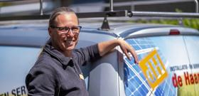 Vrouwelijke installateur kijkt lachend naar de camera en leunt tegen achterkant van werkbus met daarop logo van Installatiebedrijf De Haan.