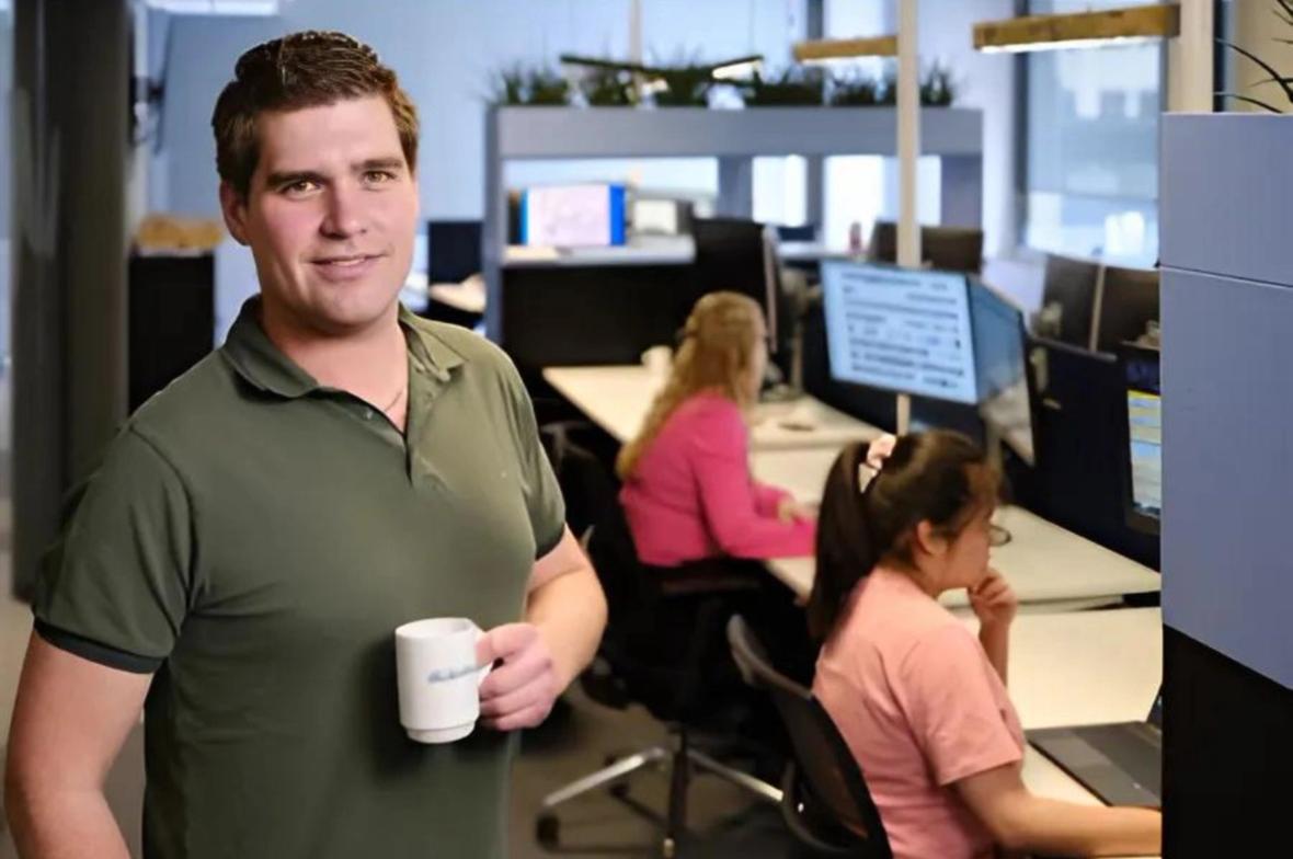 Rick de Jong Hr Adviseur van Gooiland Elektro in een groen poloshirt houdt een witte mok vast en lacht naar de camera in een kantooromgeving met twee vrouwen die op de achtergrond aan computers werken.