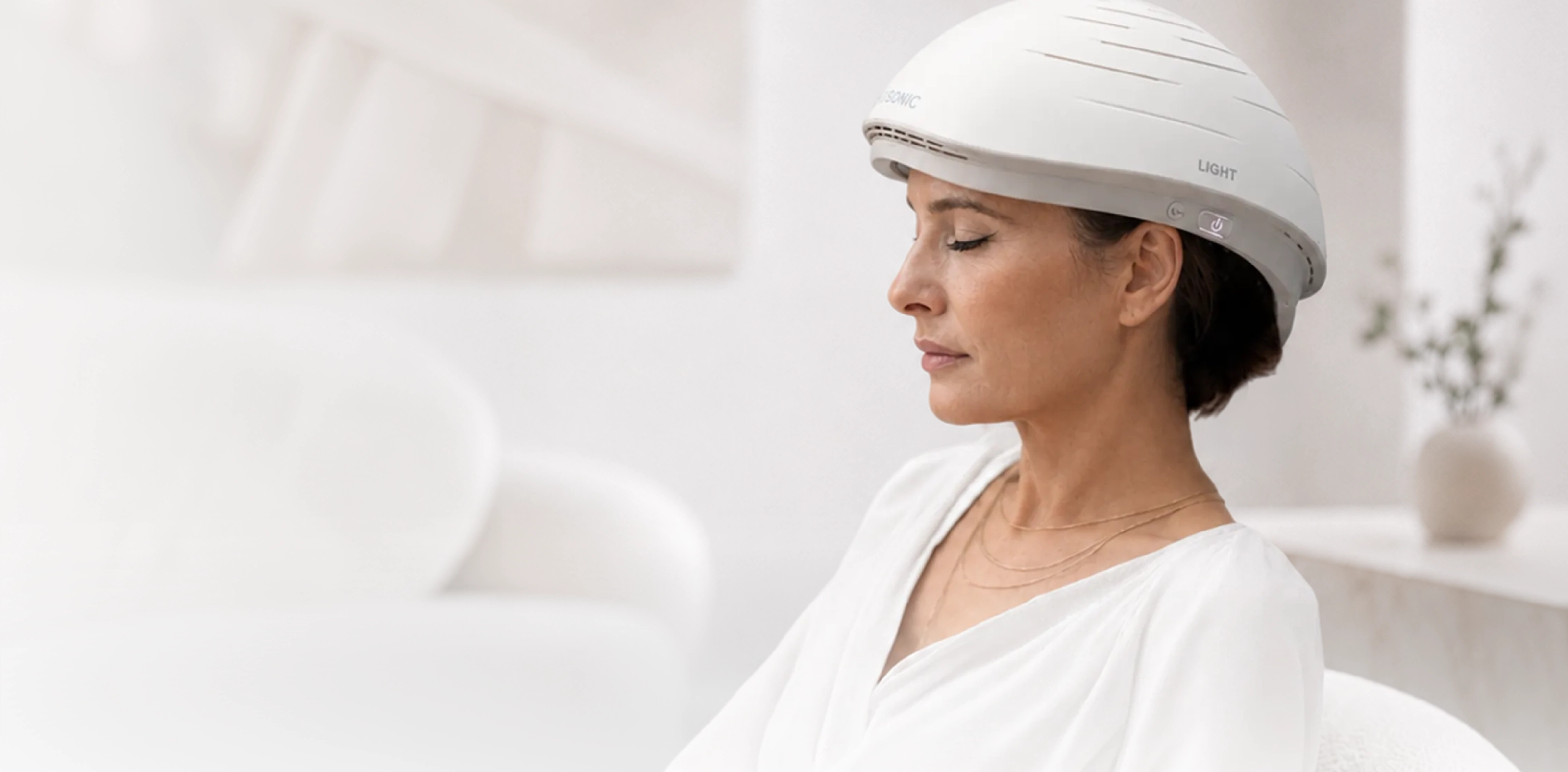 A woman sitting in a bright treatment room wearing the Neuronic LIGHT helmet with her eyes closed.