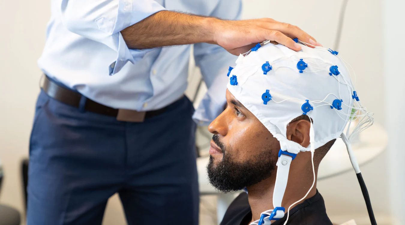 Man wearing Neuronic Helmet with blue light emitters, being fitted by a clinician in light blue shirt