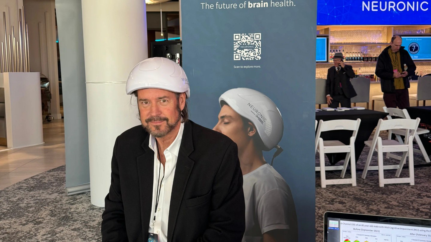 Man wearing Neuronic Helmet at product display event, with promotional backdrop showing "The future of brain health