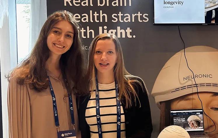 Two women wearing credential badges standing in front of a Neuronic Helmet display with tagline "Real brain health starts with light