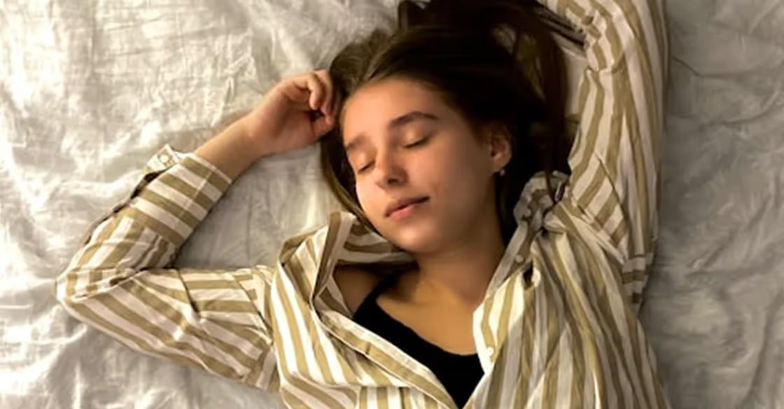 Person lying down with eyes closed, arm raised to head, wearing a striped button-up shirt over a black top