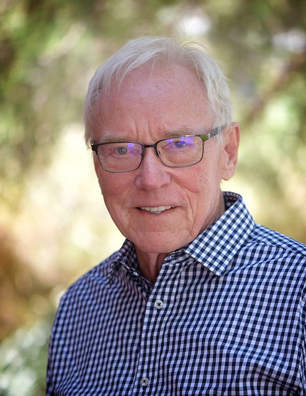 Older man wearing glasses and a blue checkered shirt, smiling at the camera outdoors