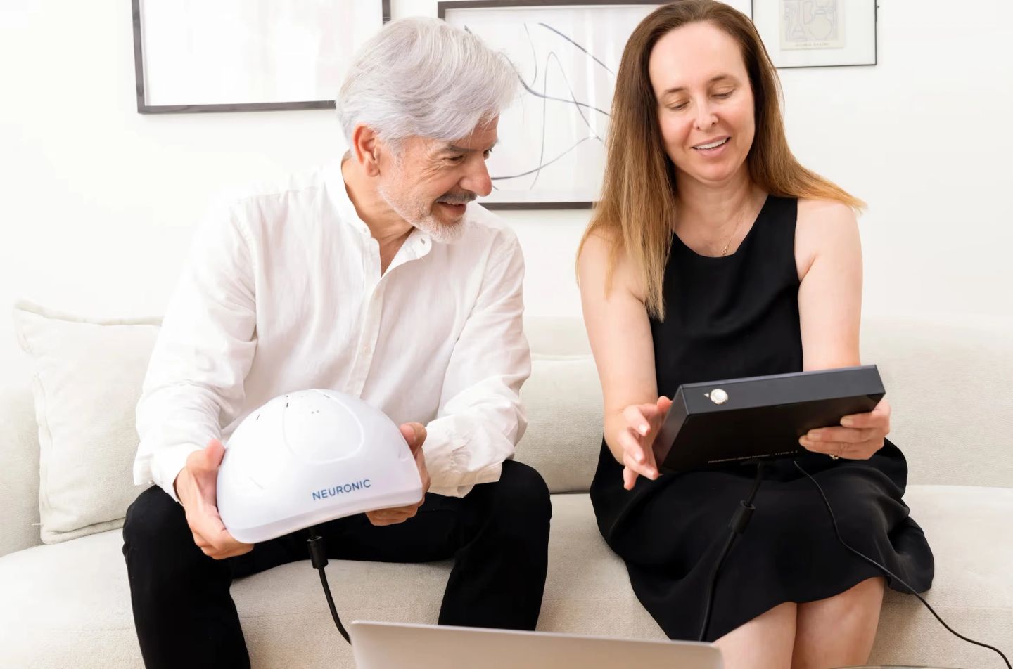 Man holding white Neuronic Helmet and woman holding control device, both looking at laptop screen while seated on couch